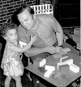 A little girl looking at the camera stands next to and reaches up to her seated dad, who leans toward her